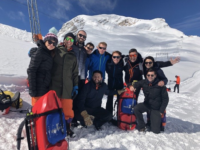 A group of nine people in winter gear posing in a snowy mountain landscape.