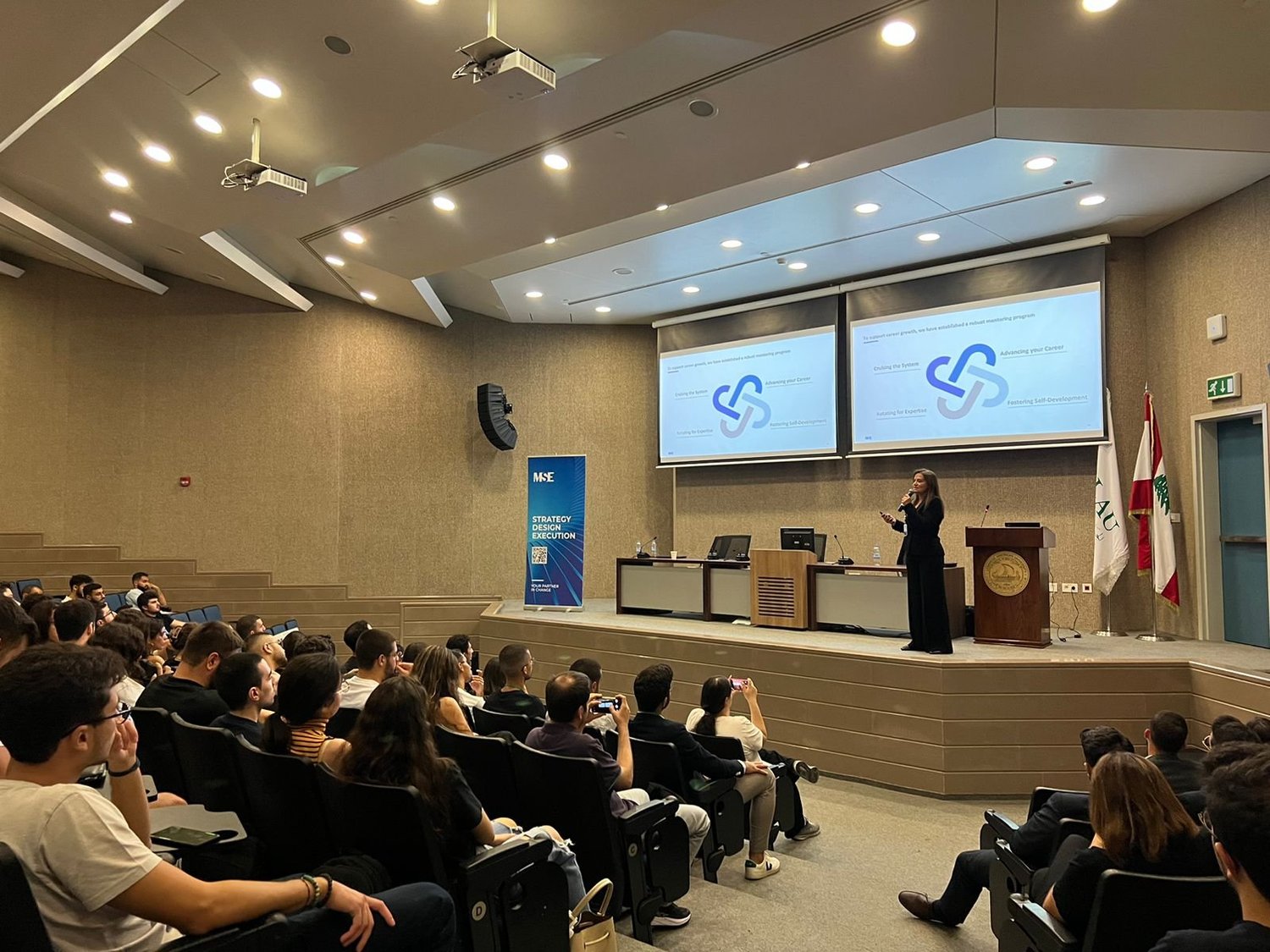 Hala Achkar Slaiby standing on stage giving a presentation to an audience in a conference hall.