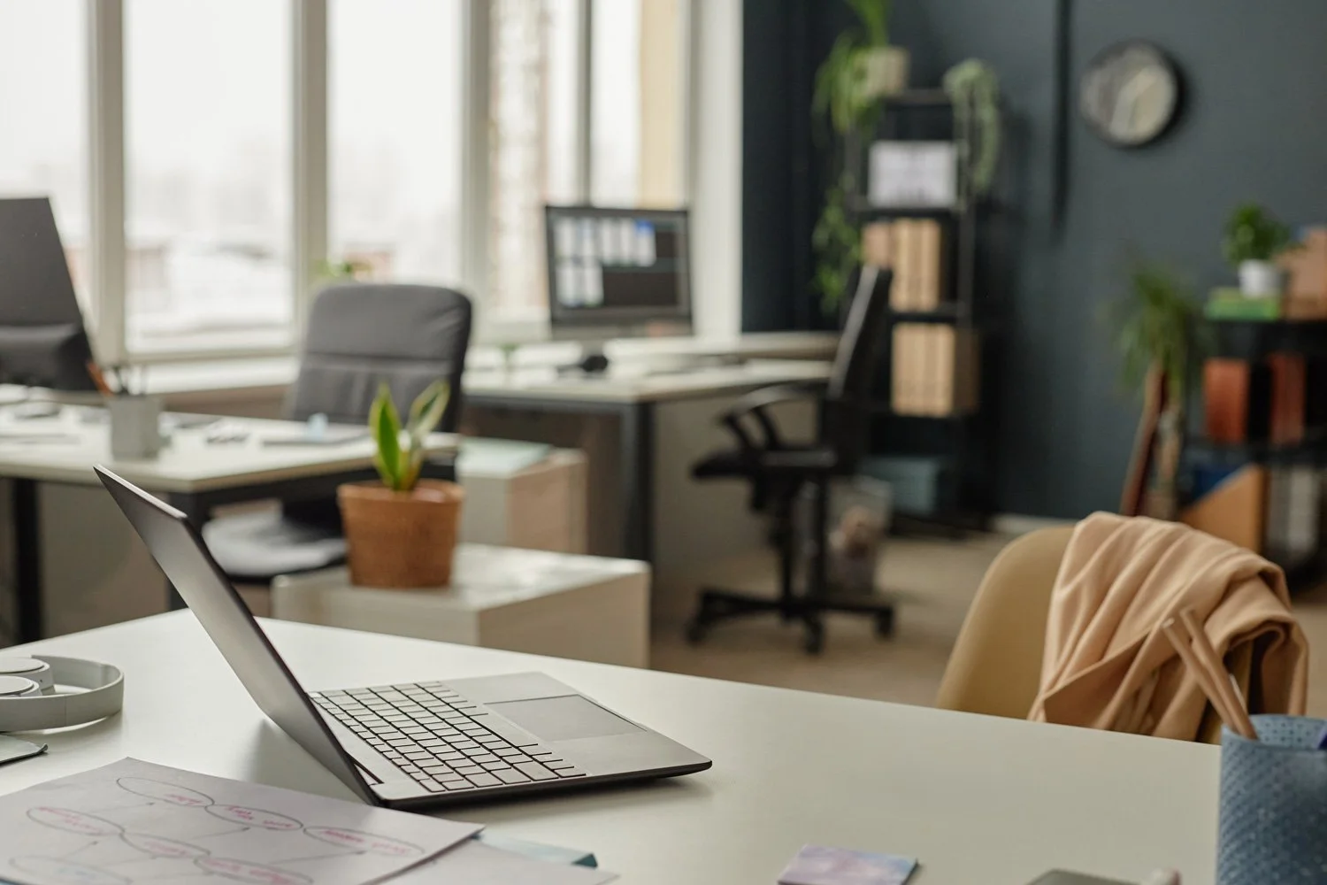 An organized office workspace with a laptop, in a bright room with large windows.